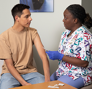 Health care provider giving teen boy injection in upper arm in exam room.