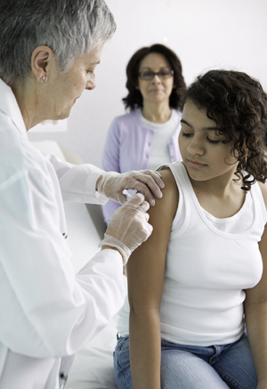 Health care provider giving shot in girl's upper arm. Woman standing in background. 