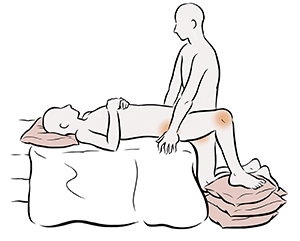 Sex position showing one person lying on bed and one person kneeling on pillows next to bed.